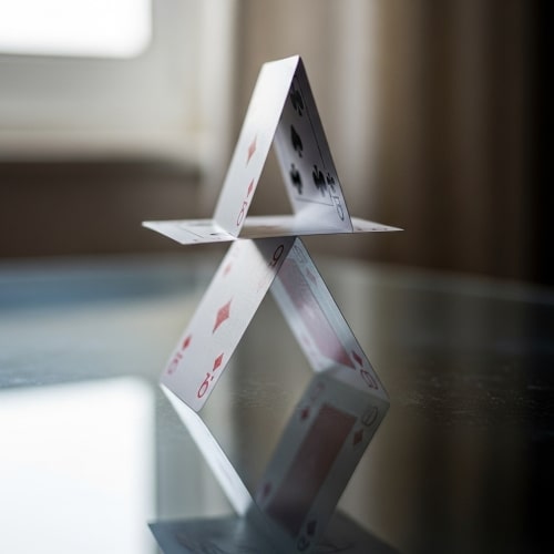 Instabiles Kartenhaus auf Glastisch – Symbol für verborgene Schwachstellen in stabil wirkenden Systemen.